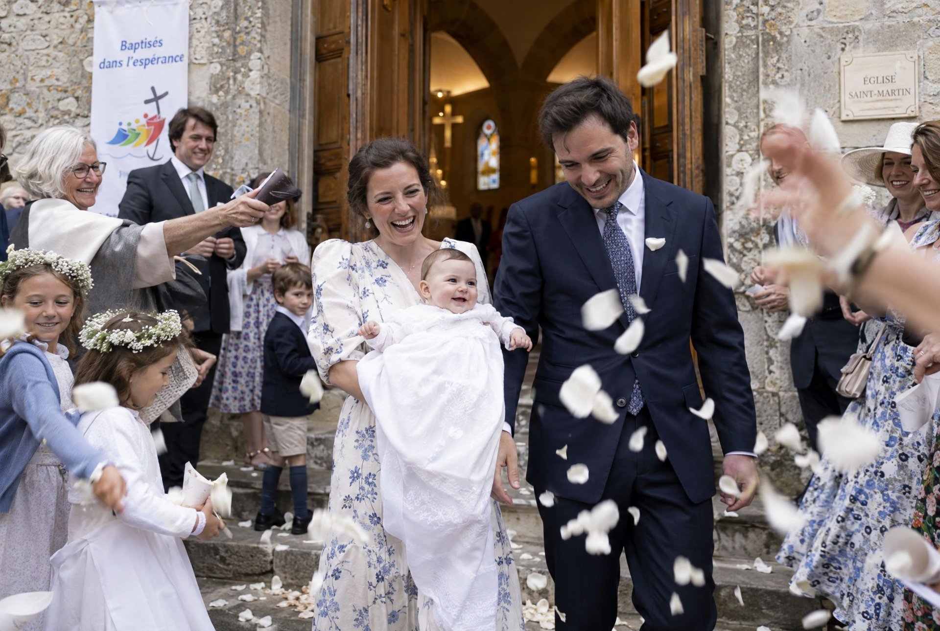 Parents portant leur bébé à la sortie de l'église sous une pluie de pétales blancs après le baptême